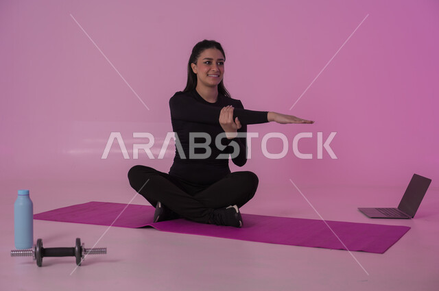 إيماءات السعادة والنشاط، أنشطة  بدنية للمحافظة على اللياقة والرشاقة، اتباع أسلوب حياة صحي باستخدام الأجهزة التقنية، رياضات لتقوية العضلات ونحت الجسم، بورتريه لشابة عربية خليجية سعودية ترتدي زي رياضي تقوم بمتابعة التمارين الرياضية عبر الحاسب المحمول، خلفية وردية