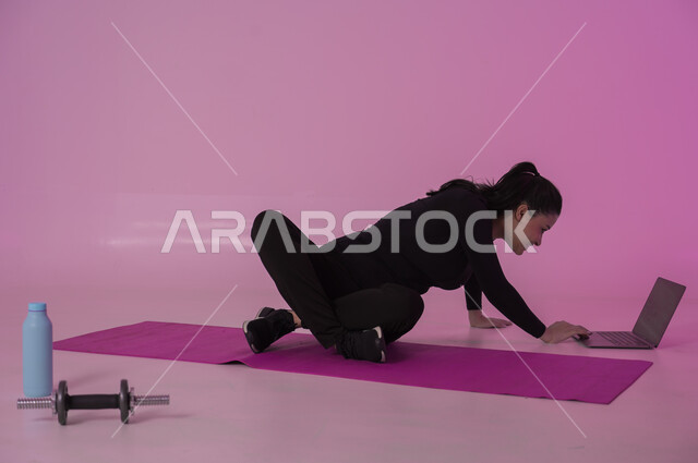 إيماءات السعادة والنشاط، أنشطة  بدنية للمحافظة على اللياقة والرشاقة، اتباع أسلوب حياة صحي باستخدام الأجهزة التقنية، رياضات لتقوية العضلات ونحت الجسم، بورتريه لشابة عربية خليجية سعودية ترتدي زي رياضي تقوم بمتابعة التمارين الرياضية عبر الحاسب المحمول، خلفية وردية