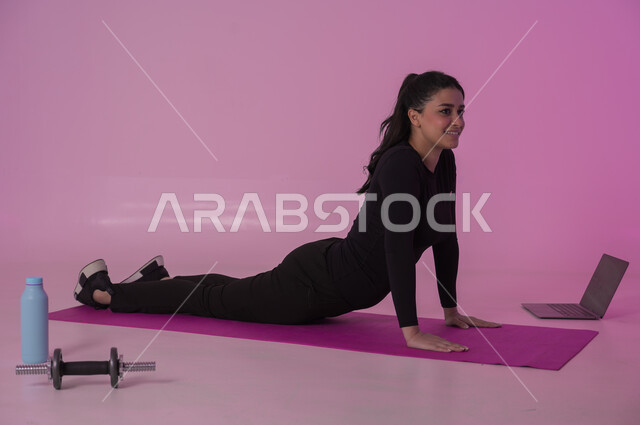 تعابير السعادة والنشاط، أنشطة  بدنية للمحافظة على اللياقة والرشاقة، اتباع أسلوب حياة صحي باستخدام الأجهزة التقنية، رياضات لتقوية العضلات ونحت الجسم، بورتريه لشابة عربية خليجية سعودية ترتدي زي رياضي تقوم بمتابعة التمارين الرياضية عبر الحاسب المحمول، خلفية وردية