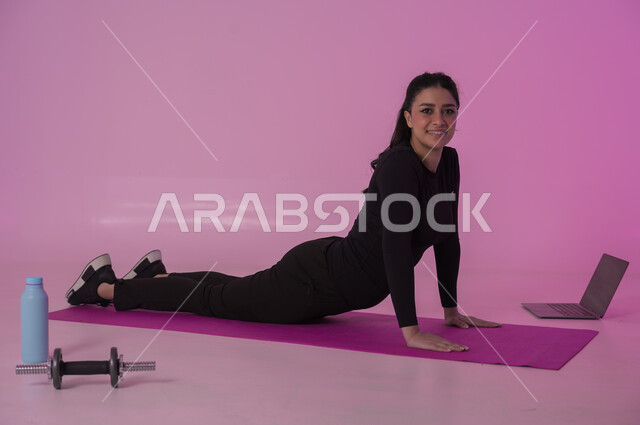 إيماءات السعادة والنشاط، أنشطة  بدنية للمحافظة على اللياقة والرشاقة، اتباع أسلوب حياة صحي باستخدام الأجهزة التقنية، رياضات لتقوية العضلات ونحت الجسم، بورتريه لشابة عربية خليجية سعودية ترتدي زي رياضي تقوم بمتابعة التمارين الرياضية عبر الحاسب المحمول، خلفية وردية