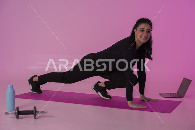 تعابير السعادة والنشاط، أنشطة  بدنية للمحافظة على اللياقة والرشاقة، اتباع أسلوب حياة صحي باستخدام الأجهزة التقنية، رياضات لتقوية العضلات ونحت الجسم، بورتريه لشابة عربية خليجية سعودية ترتدي زي رياضي تقوم بمتابعة التمارين الرياضية عبر الحاسب المحمول، خلفية وردية