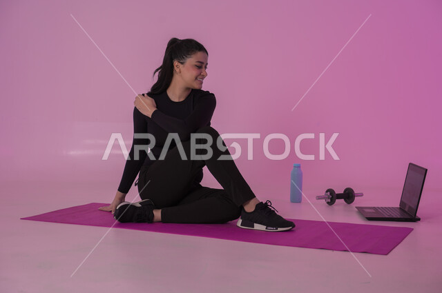  نشاط بدني للمحافظة على اللياقة والرشاقة، اتباع أسلوب حياة صحي باستخدام الأجهزة التقنية، رياضات لتقوية العضلات ونحت الجسم، إيماءات السعادة والنشاط، بورتريه لشابة عربية خليجية سعودية ترتدي زي رياضي تقوم بمتابعة التمارين الرياضية عبر الحاسب المحمول، خلفية وردية