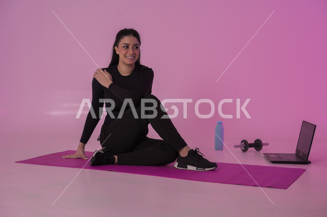  نشاط بدني للمحافظة على اللياقة والرشاقة، اتباع أسلوب حياة صحي باستخدام الأجهزة التقنية، رياضات لتقوية العضلات ونحت الجسم، إيماءات السعادة والنشاط، بورتريه لشابة عربية خليجية سعودية ترتدي زي رياضي تقوم بمتابعة التمارين الرياضية عبر الحاسب المحمول، خلفية وردية