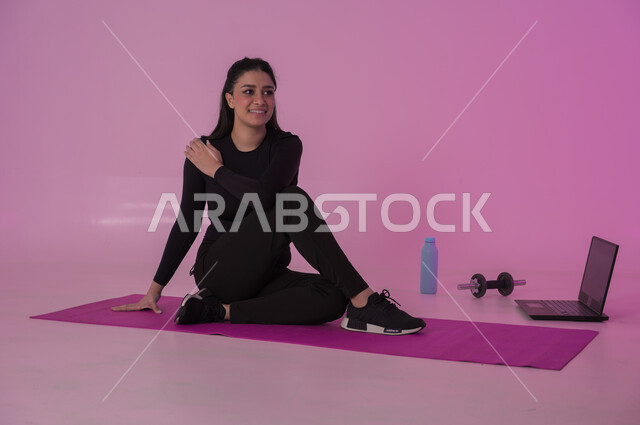  نشاط بدني للمحافظة على اللياقة والرشاقة، اتباع أسلوب حياة صحي باستخدام الأجهزة التقنية، رياضات لتقوية العضلات ونحت الجسم، إيماءات السعادة والنشاط، بورتريه لشابة عربية خليجية سعودية ترتدي زي رياضي تقوم بمتابعة التمارين الرياضية عبر الحاسب المحمول، خلفية وردية