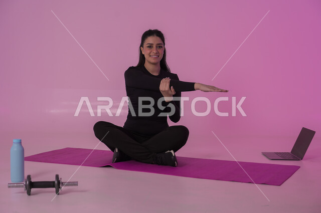 إيماءات السعادة والنشاط، أنشطة  بدنية للمحافظة على اللياقة والرشاقة، اتباع أسلوب حياة صحي باستخدام الأجهزة التقنية، رياضات لتقوية العضلات ونحت الجسم، بورتريه لشابة عربية خليجية سعودية ترتدي زي رياضي تقوم بمتابعة التمارين الرياضية عبر الحاسب المحمول، خلفية وردية