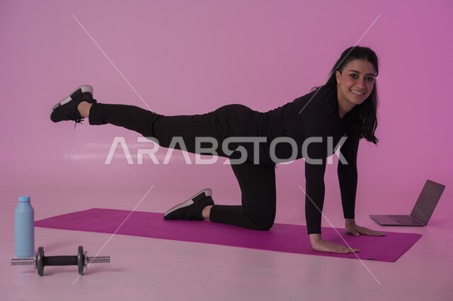 تعابير السعادة والنشاط، أنشطة  بدنية للمحافظة على اللياقة والرشاقة، اتباع أسلوب حياة صحي باستخدام الأجهزة التقنية، رياضات لتقوية العضلات ونحت الجسم، بورتريه لشابة عربية خليجية سعودية ترتدي زي رياضي تقوم بمتابعة التمارين الرياضية عبر الحاسب المحمول، خلفية وردية
