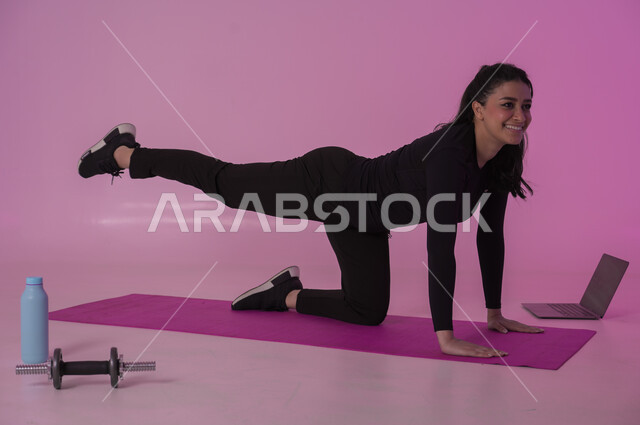 تعابير السعادة والنشاط، أنشطة  بدنية للمحافظة على اللياقة والرشاقة، اتباع أسلوب حياة صحي باستخدام الأجهزة التقنية، رياضات لتقوية العضلات ونحت الجسم، بورتريه لشابة عربية خليجية سعودية ترتدي زي رياضي تقوم بمتابعة التمارين الرياضية عبر الحاسب المحمول، خلفية وردية