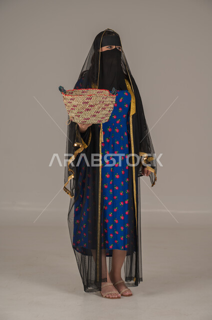 Traditional folk crafts, the anniversary of the founding of the first Saudi state, February 22, wearing the costumes of the central region on national occasions and holidays, a portrait of a Saudi Gulf Arab woman wearing a burqa and a shayla carrying a straw basket in her hand, the day of our beginnings 1727 AD, a full body image, a gray background