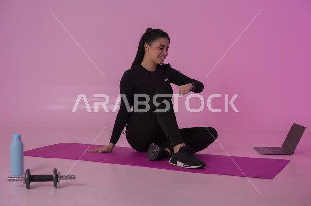 أنشطة  بدنية للمحافظة على اللياقة والرشاقة، إيماءات السعادة والنشاط، اتباع أسلوب حياة صحي باستخدام الأجهزة التقنية، رياضات لتقوية العضلات ونحت الجسم، بورتريه لشابة عربية خليجية سعودية ترتدي زي رياضي تقوم بمتابعة التمارين الرياضية عبر الحاسب المحمول، خلفية وردية