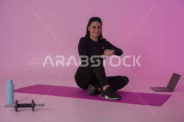 أنشطة  بدنية للمحافظة على اللياقة والرشاقة، إيماءات السعادة والنشاط، اتباع أسلوب حياة صحي باستخدام الأجهزة التقنية، رياضات لتقوية العضلات ونحت الجسم، بورتريه لشابة عربية خليجية سعودية ترتدي زي رياضي تقوم بمتابعة التمارين الرياضية عبر الحاسب المحمول، خلفية وردية