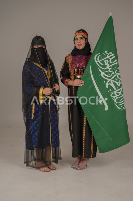  ذكرى تأسيس الدولة السعودية الاولى 22 فبراير، تعابير الفخر والاعتزاز،ارتداء أزياء المنطقة الوسطى،الاحتفال بالأعياد الوطنية، بورتريه لامرأتين عربيتين خليجيتين سعوديتين ترتديان زي يوم التأسيس يحملان علم المملكة، يوم بدينا ١٧٢٧م, الجسم الكامل بالطول، خلفية رمادية