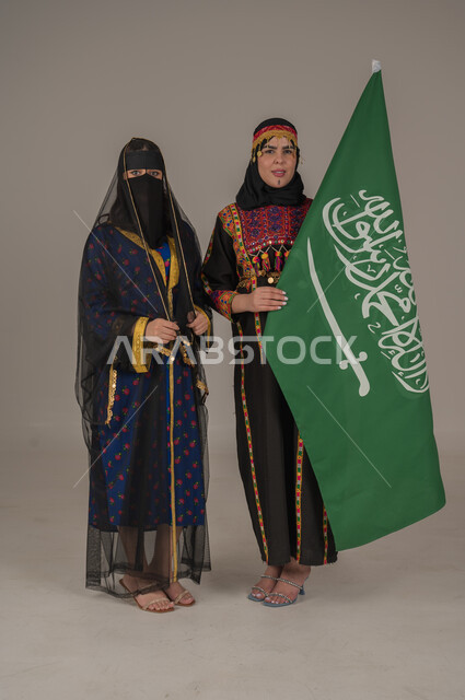 Anniversary of the founding of the first Saudi state, February 22, expressions of pride and honor, wearing the costumes of the central region, celebrating national holidays, portrait of two Saudi Arabian Gulf women wearing the Founding Day costume carrying the flag of the Kingdom, the day we began 1727 AD, full body length, gray background