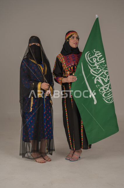 Anniversary of the founding of the first Saudi state, February 22, expressions of pride and honor, wearing the costumes of the central region, celebrating national holidays, portrait of two Saudi Arabian Gulf women wearing the Founding Day costume carrying the flag of the Kingdom, the day we began 1727 AD, full body length, gray background