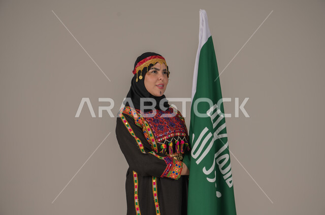 الاحتفال بالمناسبات الوطنية, يوم بدينا ١٧٢٧م، بورتريه لإمرأة عربيه سعودية خليجية ترتدي الهامة والشيلة والكرتة تحمل بيدها علم المملكة، ذكرى تأسيس الدولة السعودية الأولى، يوم العلم السعودي 11 مارس, إرتداء أزياء المنطقة الوسطى, الجسم الكامل بالطول، خلفيه رمادية
