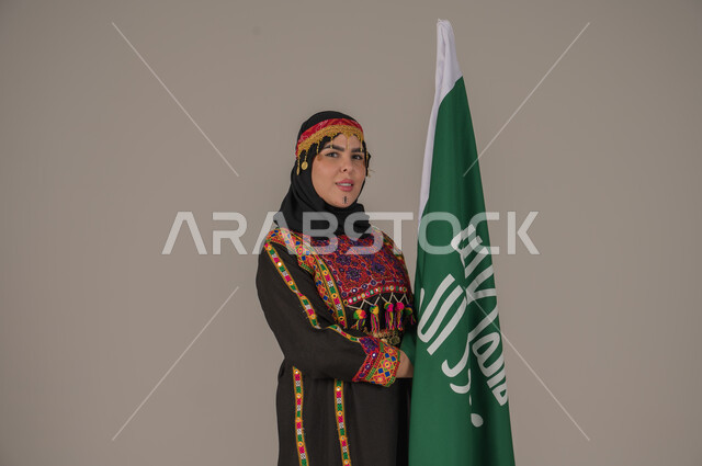 Saudi Flag Day March 11, Celebrating National Occasions, Expressions of Pride and Honor, The Day We Began 1727 AD, Portrait of a Saudi Arabian Gulf Woman Wearing a Hat, Sheila and Kurta Holding the Flag of the Founding Day, Anniversary of the Founding of the First Saudi State, Wearing the Costumes of the Central Region, Gray Background