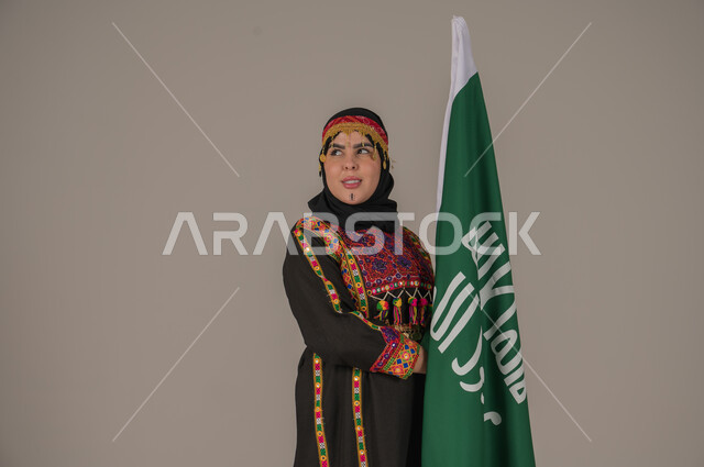Saudi Flag Day March 11, Celebrating National Occasions, Expressions of Pride and Honor, The Day We Began 1727 AD, Portrait of a Saudi Arabian Gulf Woman Wearing a Hat, Sheila and Kurta Holding the Flag of the Founding Day, Anniversary of the Founding of the First Saudi State, Wearing the Costumes of the Central Region, Gray Background