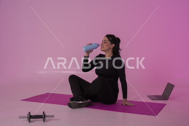 أهمية شرب الماء النقي للمحافظة على بشرة نضرة، أنشطة  بدنية للمحافظة على اللياقة والرشاقة،  اتباع أسلوب حياة صحي باستخدام الأجهزة التقنية، بورتريه لشابة عربية خليجية سعودية ترتدي زي رياضي تقوم بمتابعة التمارين الرياضية عبر الحاسب المحمول، خلفية وردية