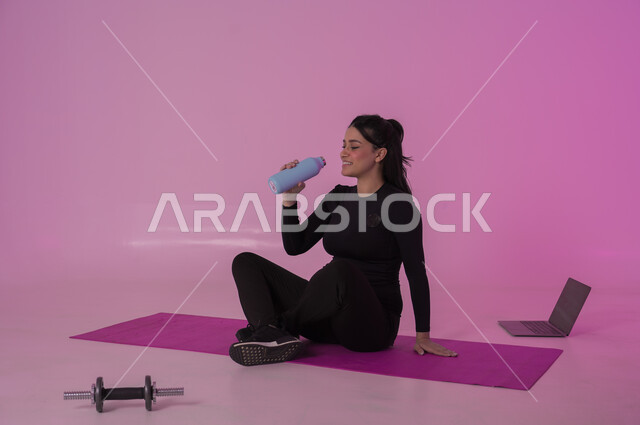أهمية شرب الماء النقي للمحافظة على بشرة نضرة، أنشطة  بدنية للمحافظة على اللياقة والرشاقة،  اتباع أسلوب حياة صحي باستخدام الأجهزة التقنية، بورتريه لشابة عربية خليجية سعودية ترتدي زي رياضي تقوم بمتابعة التمارين الرياضية عبر الحاسب المحمول، خلفية وردية