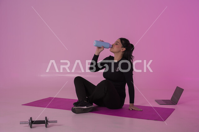 أهمية شرب الماء النقي للمحافظة على بشرة نضرة، أنشطة  بدنية للمحافظة على اللياقة والرشاقة،  اتباع أسلوب حياة صحي باستخدام الأجهزة التقنية، بورتريه لشابة عربية خليجية سعودية ترتدي زي رياضي تقوم بمتابعة التمارين الرياضية عبر الحاسب المحمول، خلفية وردية