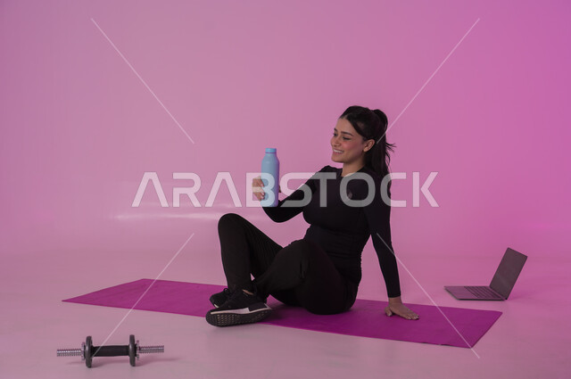 أهمية شرب الماء النقي للمحافظة على بشرة نضرة، أنشطة  بدنية للمحافظة على اللياقة والرشاقة،  اتباع أسلوب حياة صحي باستخدام الأجهزة التقنية، بورتريه لشابة عربية خليجية سعودية ترتدي زي رياضي تقوم بمتابعة التمارين الرياضية عبر الحاسب المحمول، خلفية وردية