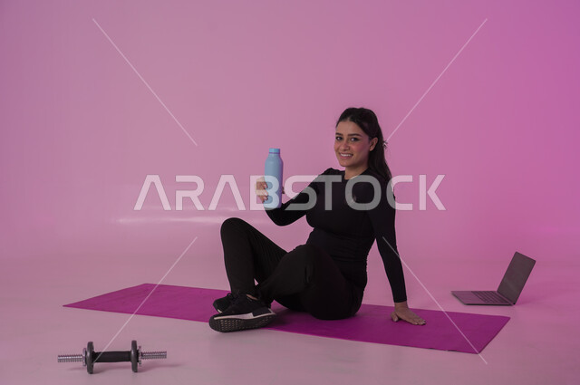 أهمية شرب الماء النقي للمحافظة على بشرة نضرة، أنشطة  بدنية للمحافظة على اللياقة والرشاقة،  اتباع أسلوب حياة صحي باستخدام الأجهزة التقنية، بورتريه لشابة عربية خليجية سعودية ترتدي زي رياضي تقوم بمتابعة التمارين الرياضية عبر الحاسب المحمول، خلفية وردية