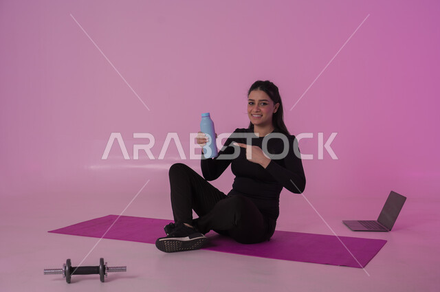 أهمية شرب الماء النقي للمحافظة على بشرة نضرة، أنشطة  بدنية للمحافظة على اللياقة والرشاقة،  اتباع أسلوب حياة صحي باستخدام الأجهزة التقنية، بورتريه لشابة عربية خليجية سعودية ترتدي زي رياضي تقوم بمتابعة التمارين الرياضية عبر الحاسب المحمول، خلفية وردية