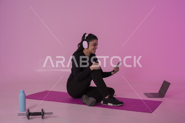 الاستماع إلى الموسيقا المفضلة والاستمتاع بها، أنشطة  بدنية للمحافظة على اللياقة والرشاقة،  اتباع أسلوب حياة صحي باستخدام الأجهزة التقنية، بورتريه لشابة عربية خليجية سعودية ترتدي زي رياضي تضع سماعة الهيدفون تقوم بمتابعة التمارين الرياضية عبر الحاسب المحمول، خلفية وردية