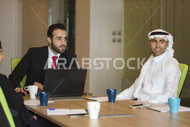 اجتماع اداري مع مجموعة من الأشخاص السعوديين الخليجيين مع رجل عربي ببدلة يتناقشون في غرفة الاجتماعات، إستخدام جهاز الكمبيوتر المحمول، شركة سعودية، شركاء العمل ، بيئة العمل، وظائف ومهن مكتبية، إجتماع عمل في مقر شركة، توقيع العقود