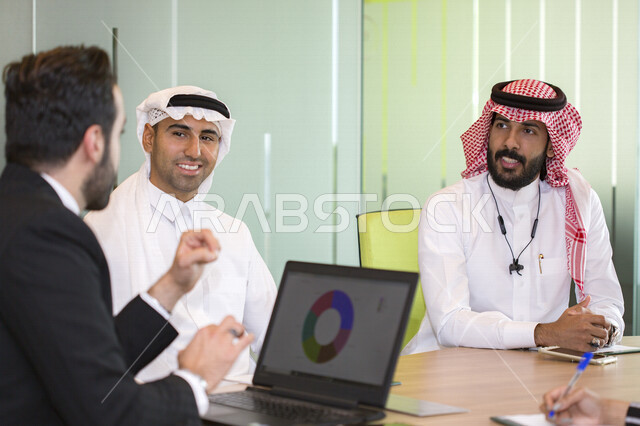 اجتماع اداري مع مجموعة من الأشخاص السعوديين الخليجيين مع رجل عربي ببدلة يتناقشون في غرفة الاجتماعات، إستخدام جهاز الكمبيوتر المحمول، شركة سعودية، شركاء العمل ، بيئة العمل، وظائف ومهن مكتبية، إجتماع عمل في مقر شركة، توقيع العقود