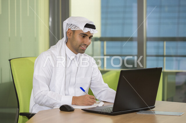 رجل اعمال عربي سعودي خليجي يعمل في مقر الشركة ، التدوين ومراجعة الملاحظات ، إستخدام الكمبيوتر المحمول لإنجاز المهام، موظف سعودي خليجي يرتدي الزي السعودي التقليدي ، شركة سعودية، وظيفة مكتبية، بيئة عمل ، أعمال تجارية