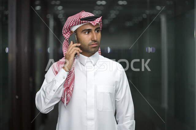 صورة مقربة لرجل اعمال عربي سعودي خليجي ، يتحدث بالهاتف المحمول في داخل مقر الشركة ، يرتدي الزي السعودي التقليدي ، شركة سعودية ، وظيفة مكتبية ، بيئة عمل ، مكالمة صوتية على الهاتف الخلوي