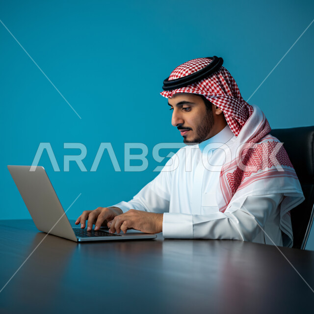 إدارة مهام العمل اليومية في مقر الشركة، مهن ووظائف سعودية، استخدام الأجهزة التقنية الحديثة، بورتريه مقرب لرجل عربي خليجي سعودي يرتدي الثوب التقليدي والشماغ يجلس في مكتبه يعمل على جهاز الحاسوب المحمول، تطوير الخطط والاسترتيجيات، خلفية زرقاء