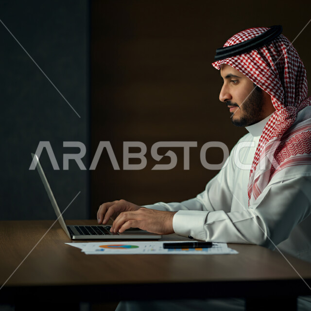 إدارة مهام العمل اليومية في مقر الشركة، مهن ووظائف سعودية، استخدام الأجهزة التقنية الحديثة، رجل عربي خليجي سعودي يرتدي الثوب التقليدي والشماغ يجلس في مكتبه يعمل على جهاز الحاسوب المحمول، تطوير الخطط والاسترتيجيات