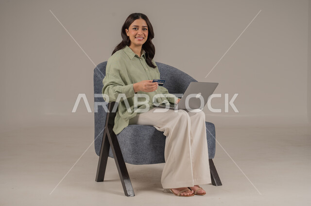 Online shopping and payment, using credit card in online shopping, portrait of a Saudi Arabian Gulf woman wearing casual clothes sitting on a chair holding a bank card and browsing banking applications on a laptop, beige background