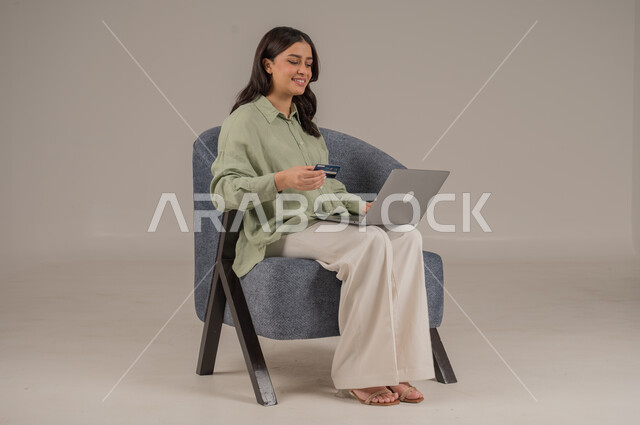 الشراء والدفع عن بعد عبر الاونلاين, استخدام بطاقة الإئتمان في التسوق الالكتروني، بورتريه  لامراة عربية سعودية خليجية ترتدي ثياب كاجوال تجلس على كرسي يمسك البطاقة البنكية ويقوم بتصفح تطبيقات المصارف والبنوك عبر اللابتوب، خلفية بيج