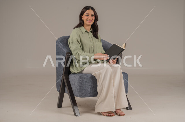 قراءة الروايات والقصص، ايماءات التركيز والاستمتاع، تمضية أوقات الفراغ بالتعلم والتثقيف الذاتي، بورتريه لامراة عربية سعودية خليجية ترتدي ثياب كاجوال تحمل بيدها كتاب, خلفية بيج 