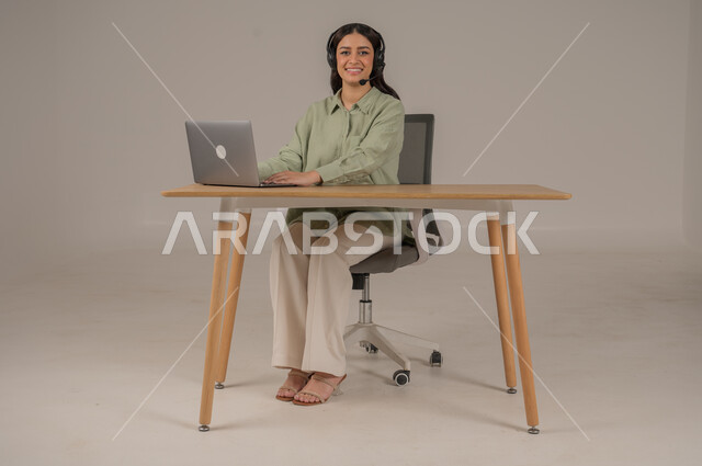 Looking at camera with gestures of pleasure, using modern and advanced technologies, responding and answering inquiries, working in customer service, portrait of a Saudi Arabian Gulf woman wearing casual clothes sitting on a chair working on a laptop, full body photo, gray background