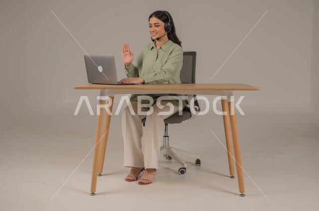 اجراء مكالمات فيديو خلال اجتماع العمل، بورتريه لإمرأة عربية خليجية سعودية ترتدي الزي الكاجوال تضع سماعات الهيدفون تعمل في مجال خدمة العملاء، الرد على الاستعلامات والاستفسارات، استخدام التقنيات الحديثة والمتطورة، صورة لكامل الجسم، خلفية رمادية