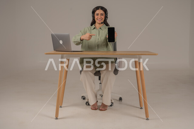 الرد والاجابة على الاستفسارات، العمل في مجال خدمة العملاء، استخدام التقنيات الحديثة، بورتريه لامراة عربية خليجية سعودية ترتدي الزي الكاجوال تجلس على الكرسي بجانب اللابتوب تشير باصبع السبابة للهاتف المحمول، عرض شاشة سوداء فارغة، صورة لكامل الجسم، خلفية رمادية
