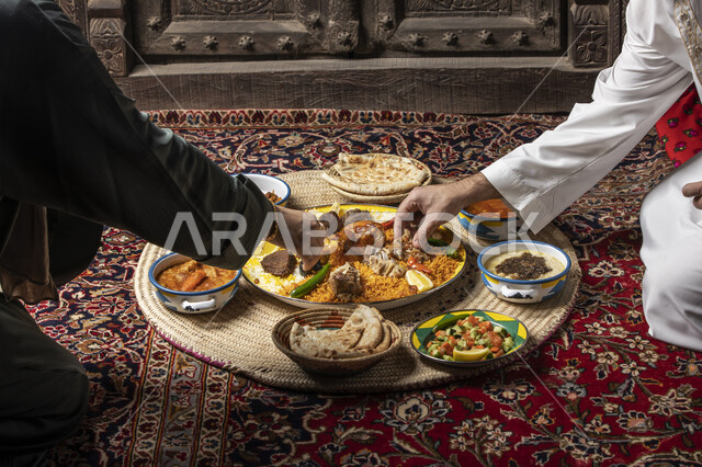 سفرة من المأكولات الشعبية التقليدية، صورة مقربة ليد زوجين عربيين خليجيين سعوديين يجلسان على الأرض يأكلان طبق الكبسة مع اللحم والخضار، المطبخ السعودي الغني بالأصناف والمقبلات المتنوعة، وصفات الطعام الصحية الشهية