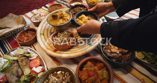 سفرة من المأكولات الشعبية التقليدية الشهية، طاولة مليئة بالوجبات المتنوعة اللذيذة، الأصناف والمقبلات الشرقية، صورة مقربة لأيدي امرأة عربية خليجية سعودية تقوم بتقديم الطعام وترتيب الأطباق، المطبخ السعودي الغني بوصفات الطهي المتميزة