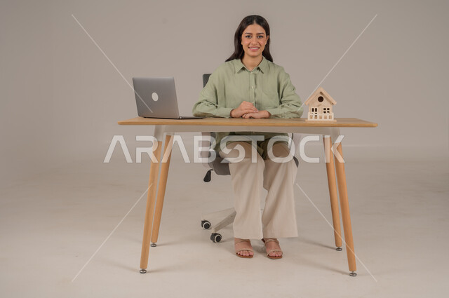 مهن ووظائف مكتبية، استخدام التقنيات الحديثة والمتطورة، الاسكان والتمليك اونلاين، بيع وشراء العقارات عبر الانترنت، ورتريه لامراة عربية خليجية سعودية ترتدي الزي الكاجوال تجلس بجانب مجسم خشبي لمنزل، صورة لكامل الجسم، خلفية رمادية