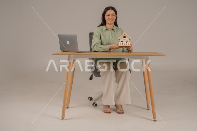 استخدام التقنيات الحديثة والمتطورة، الاسكان والتمليك اونلاين، بيع وشراء العقارات عبر الانترنت، ورتريه لامراة عربية خليجية سعودية ترتدي الزي الكاجوال تحمل بيدها مجسم خشبي لمنزل، مهن ووظائف مكتبية، صورة لكامل الجسم، خلفية رمادية