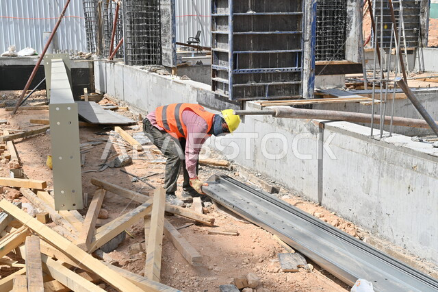 أعمال البناء وصب الخرسانات ووضع الأساسات الأولية، تنفيذ مخطط الإعمار وتشكيل الهيكل المعدني، الآلات والمعدات الثقيلة والرافعات والسقالات العالية، عامل عربي خليجي سعودي يرتدي سترة وخوذة الحماية يقوم بالعمل في مبنى قيد الإنشاء بالمملكة العربية السعودية