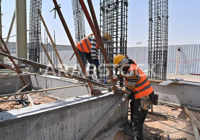 المعدات الثقيلة والرافعات والسقالات العالية المستخدمة في موقع المشروع، أعمال البناء وصب الخرسانات ووضع الأساسات الأولية، تنفيذ مخطط الإعمار وتشكيل الهيكل المعدني، عمال عربيون خليجيون سعوديون يرتدون سترة وخوذة الحماية يقومون بالعمل في مبنى قيد الإنشاء والتشييد