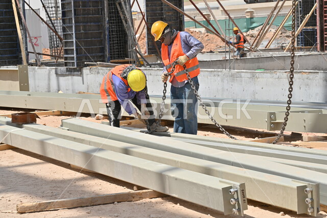 تنفيذ مخطط الإعمار وتشكيل الهيكل المعدني، المعدات الثقيلة والرافعات والسقالات العالية المستخدمة في موقع المشروع، أعمال البناء وصب الخرسانات ووضع الأساسات الأولية، عمال عربيون خليجيون سعوديون يرتدون سترة وخوذة الحماية يقومون بالعمل في مبنى قيد الإنشاء والتشييد