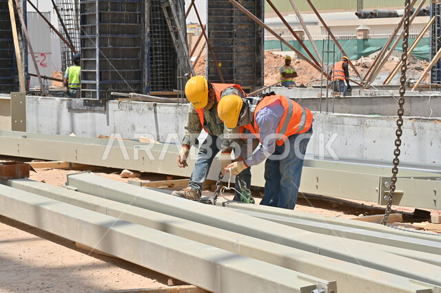 تنفيذ مخطط الإعمار وتشكيل الهيكل المعدني، المعدات الثقيلة والرافعات والسقالات العالية المستخدمة في موقع المشروع، أعمال البناء وصب الخرسانات ووضع الأساسات الأولية، عمال عربيون خليجيون سعوديون يرتدون سترة وخوذة الحماية يقومون بالعمل في مبنى قيد الإنشاء والتشييد