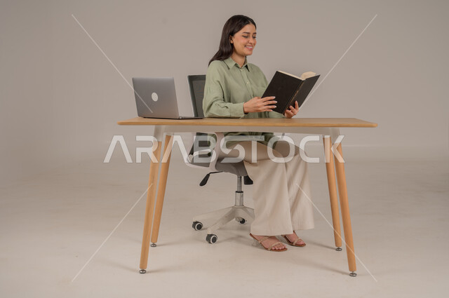 قضاء الوقت بالمطالعة والتثقيف، ممارسة الهوايات المفضلة، بورتريه لامراة عربية خليجية سعودية ترتدي الزي الكاجوال تجلس على طاولة المكتب تقوم بقراءة كتاب، استخدام التقنيات الحديثة والمتطورة، صورة لكامل الجسم، خلفية رمادية