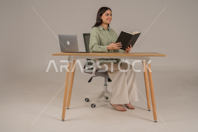 Spending time reading and educating, practicing favorite hobbies, portrait of a Saudi Arabian Gulf woman wearing casual clothes sitting at an office table reading a book, using modern and advanced technologies, full body image, gray background