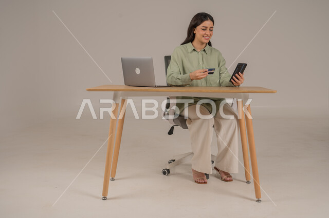 تسديد الفواتير عبر الحساب البنكي، الشراء والدفع عن طريق بطاقة الصراف الإئتمانية، بورتريه لامراة عربية خليجية سعودية ترتدي الزي الكاجوال تجلس على طاولة المكتب تحمل بيدها كرت البنك المصرفي والهاتف المحمول، صورة لكامل الجسم، خلفية رمادية